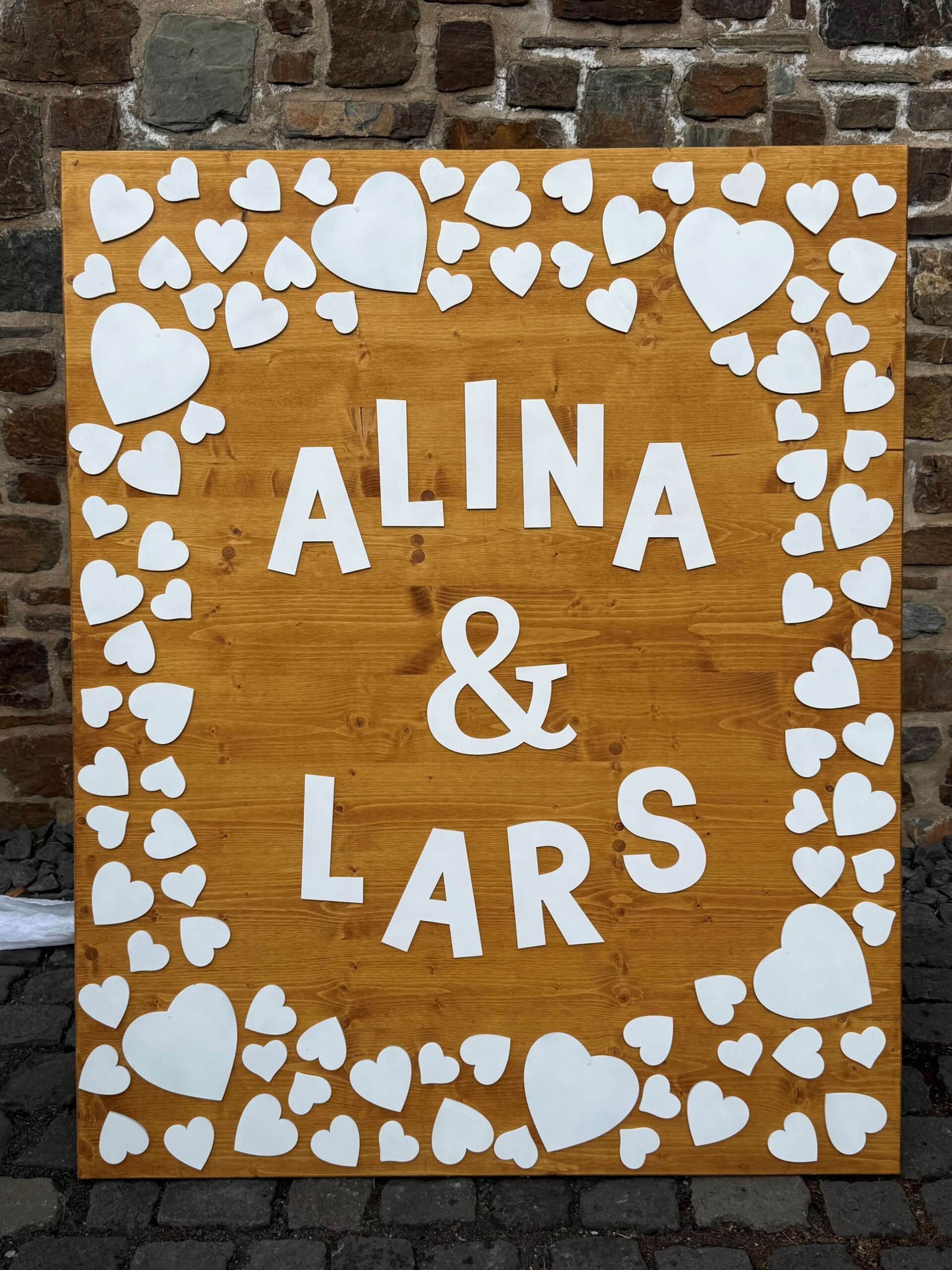 Hölzernes Hochzeitsschild mit weißen Herzen und den Namen Alina und Lars vor einer Steinwand